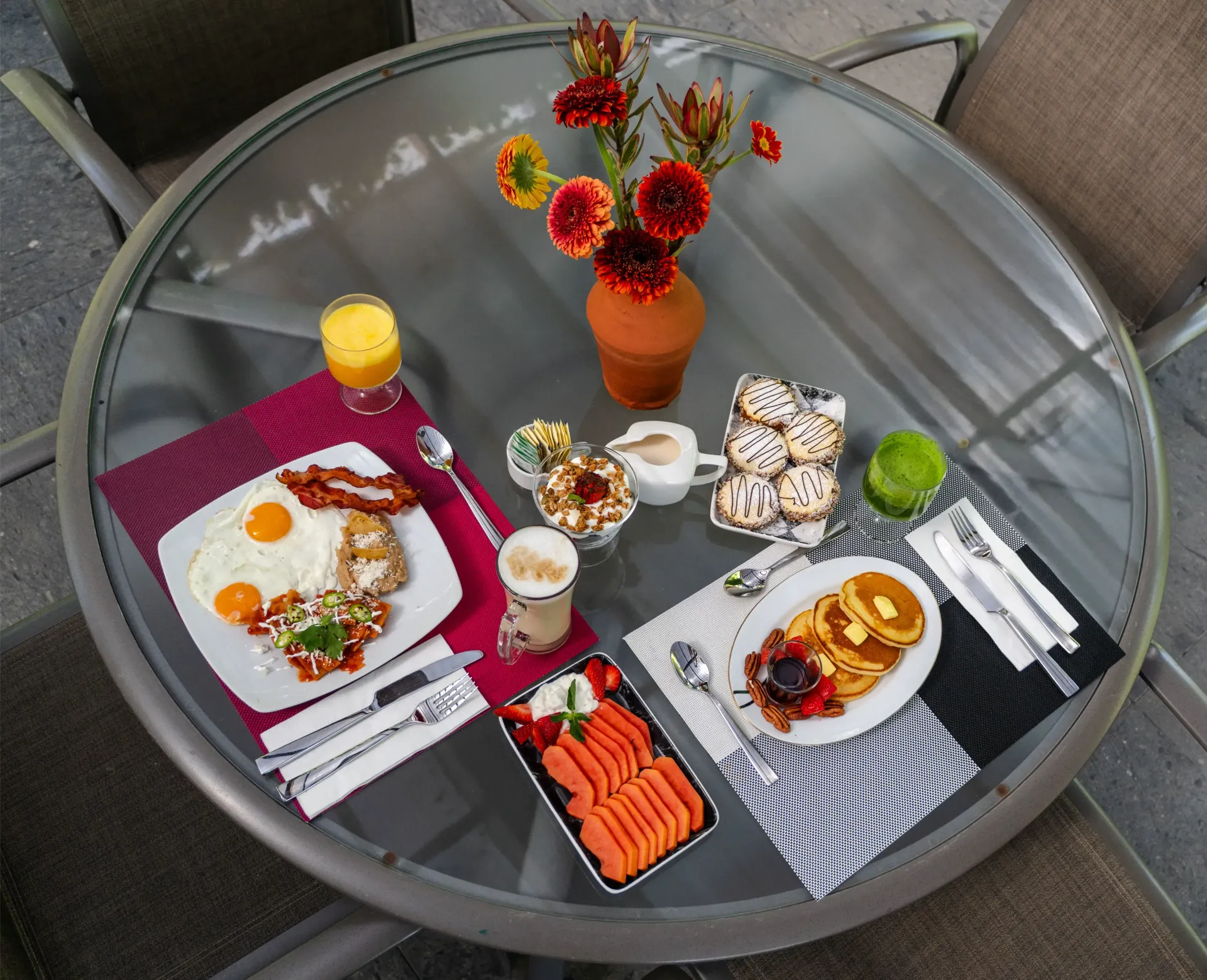 Mesa al aire libre con una selección de platos de brunch, bebidas y un jarrón con flores rojas y amarillas