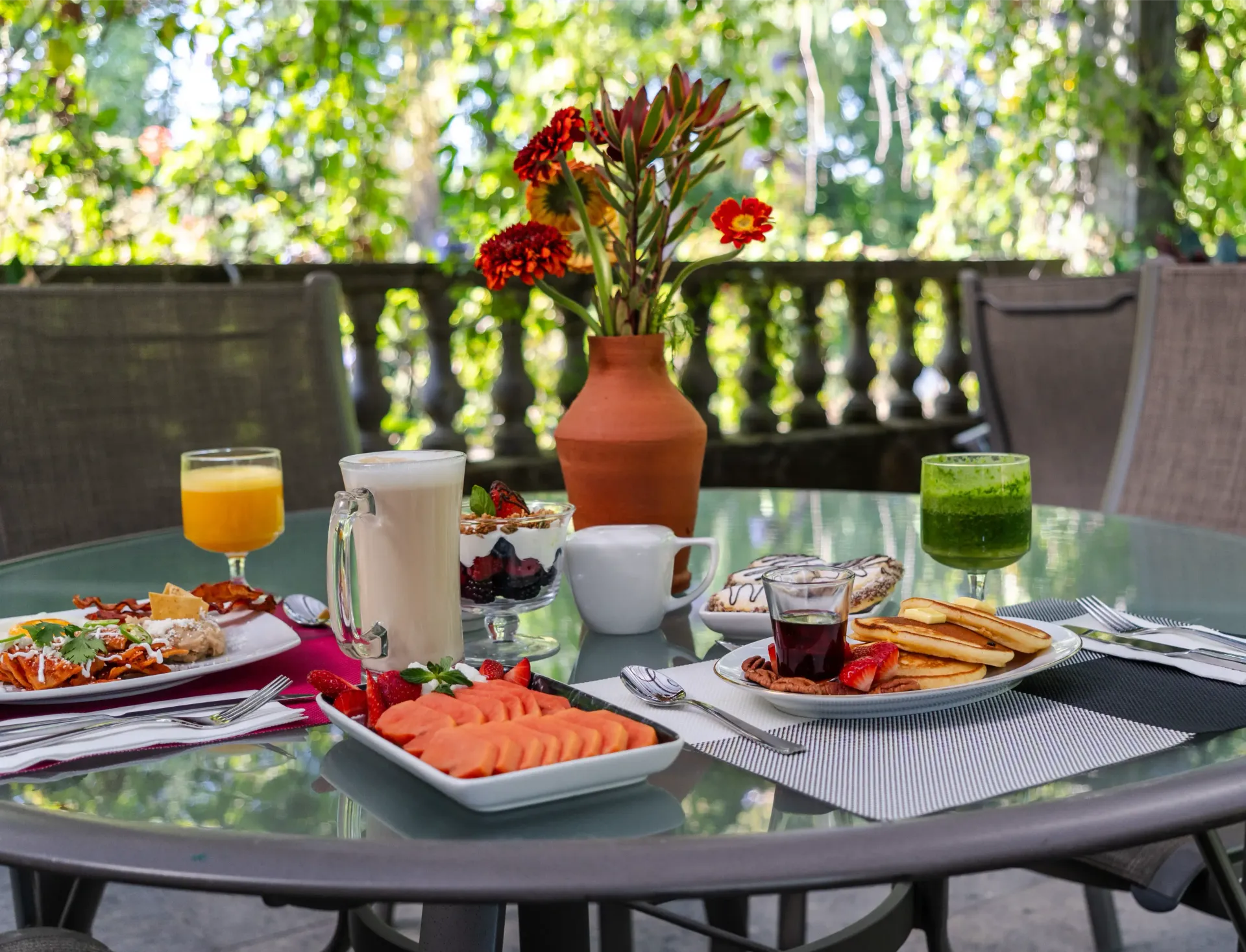 Juego de mesa de exterior con platos de desayuno, zumo de naranja, café, fruta y flores en un jarrón