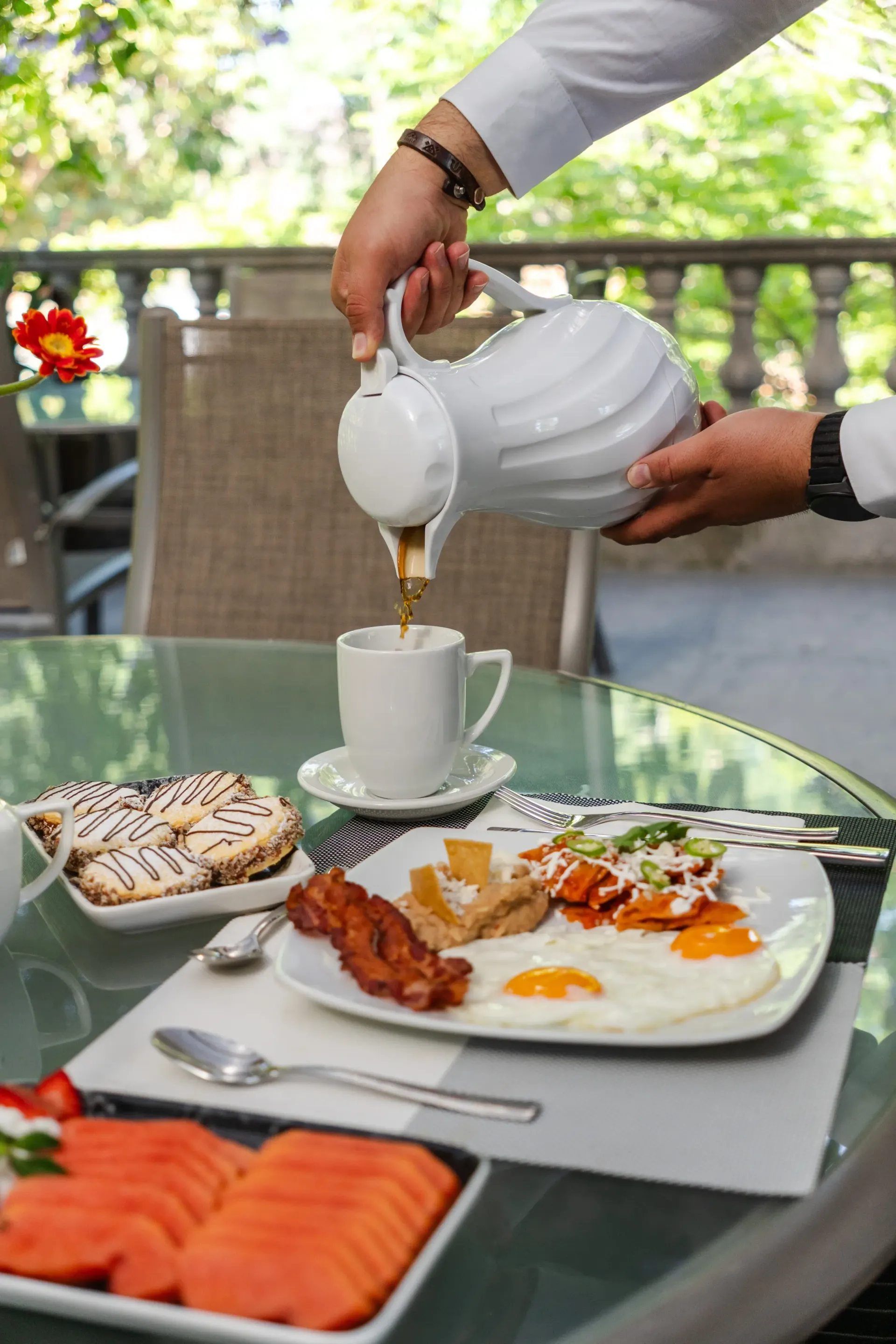 Té sirviéndose en una taza blanca, sobre una mesa al aire libre con bollería y fruta para el desayuno.