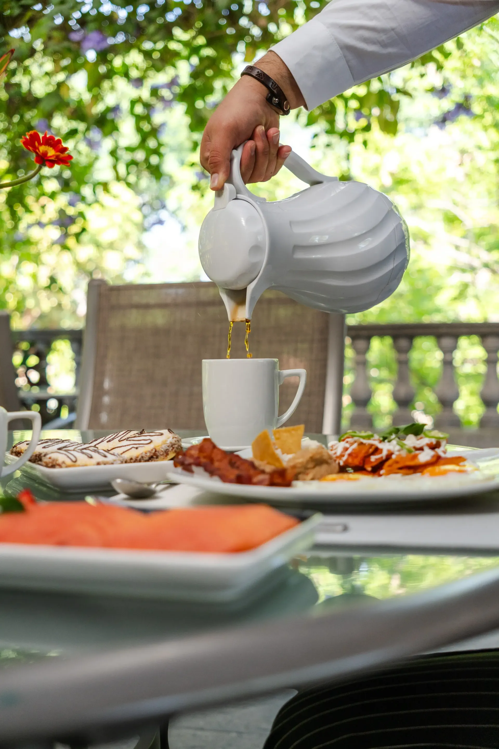 Una mano sirve té de una tetera blanca en una taza blanca, en una mesa al aire libre con comida y platos.