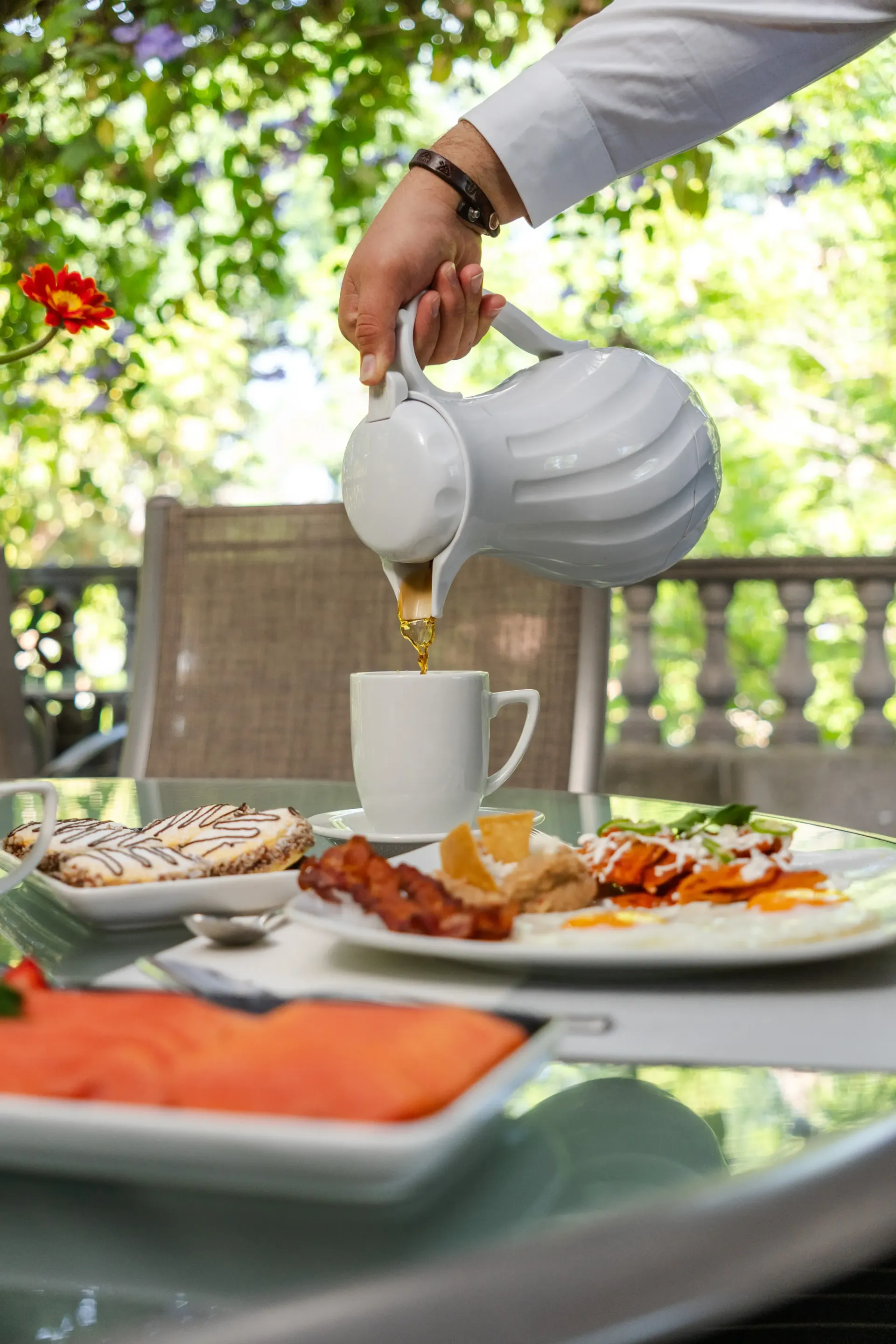 Una mano sirve té de una tetera blanca en una taza sobre una mesa al aire libre con platos de desayuno