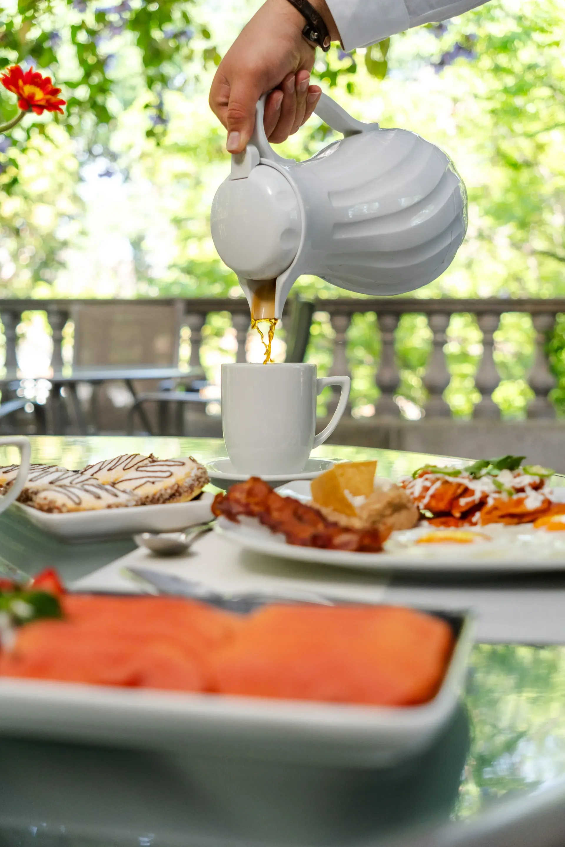 Una mano sirviendo té en una taza blanca en una mesa al aire libre con pasteles y salmón ahumado.