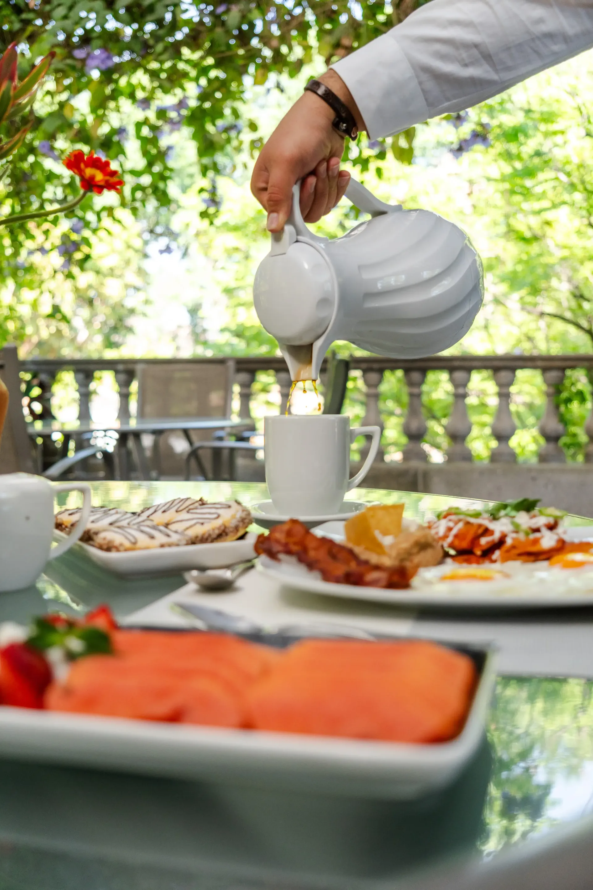 Una mano vierte leche en una taza blanca en una mesa de desayuno al aire libre con fruta y bollería