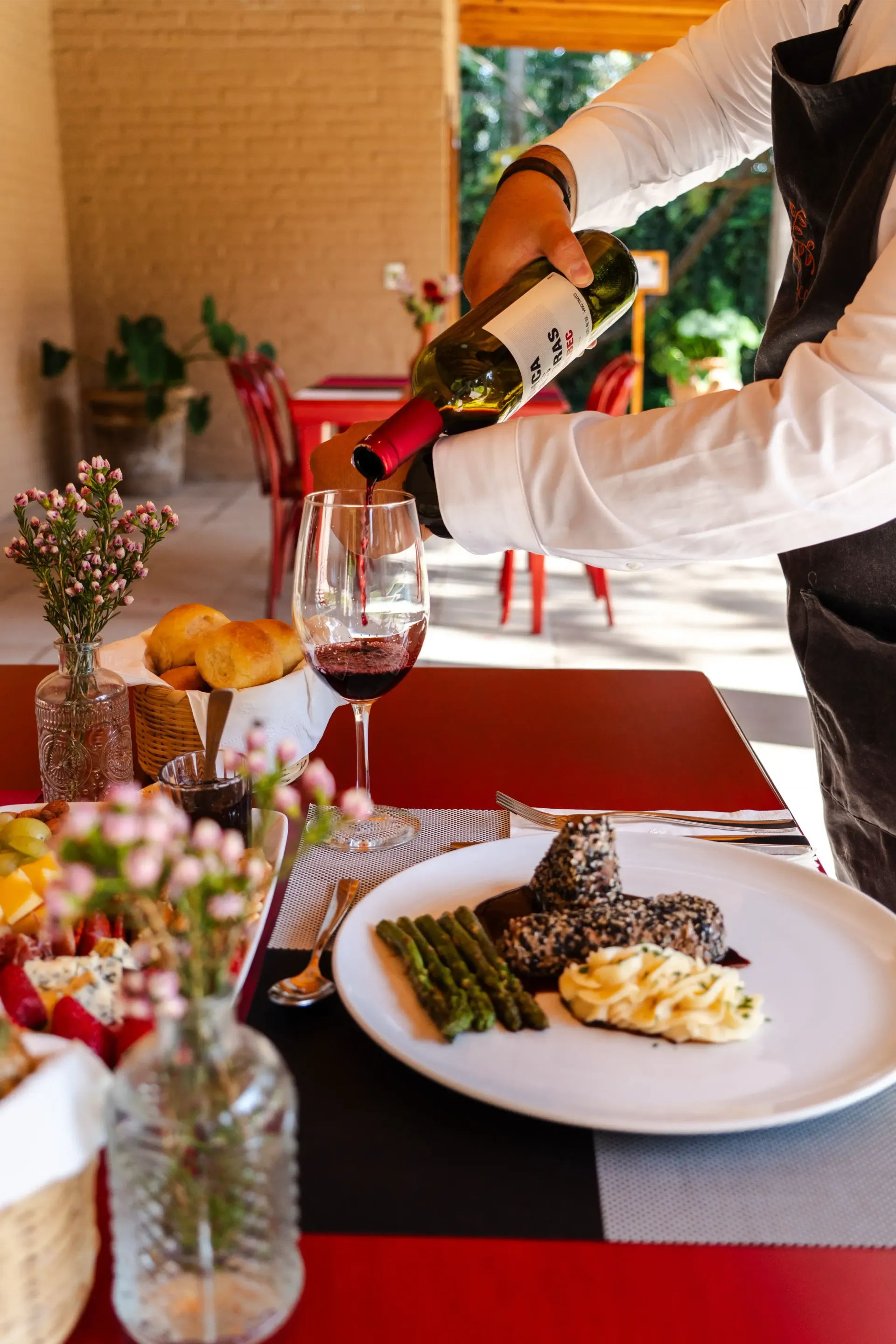 Un camarero sirviendo vino tinto en una mesa de restaurante iluminada con velas, con platos de comida y flores