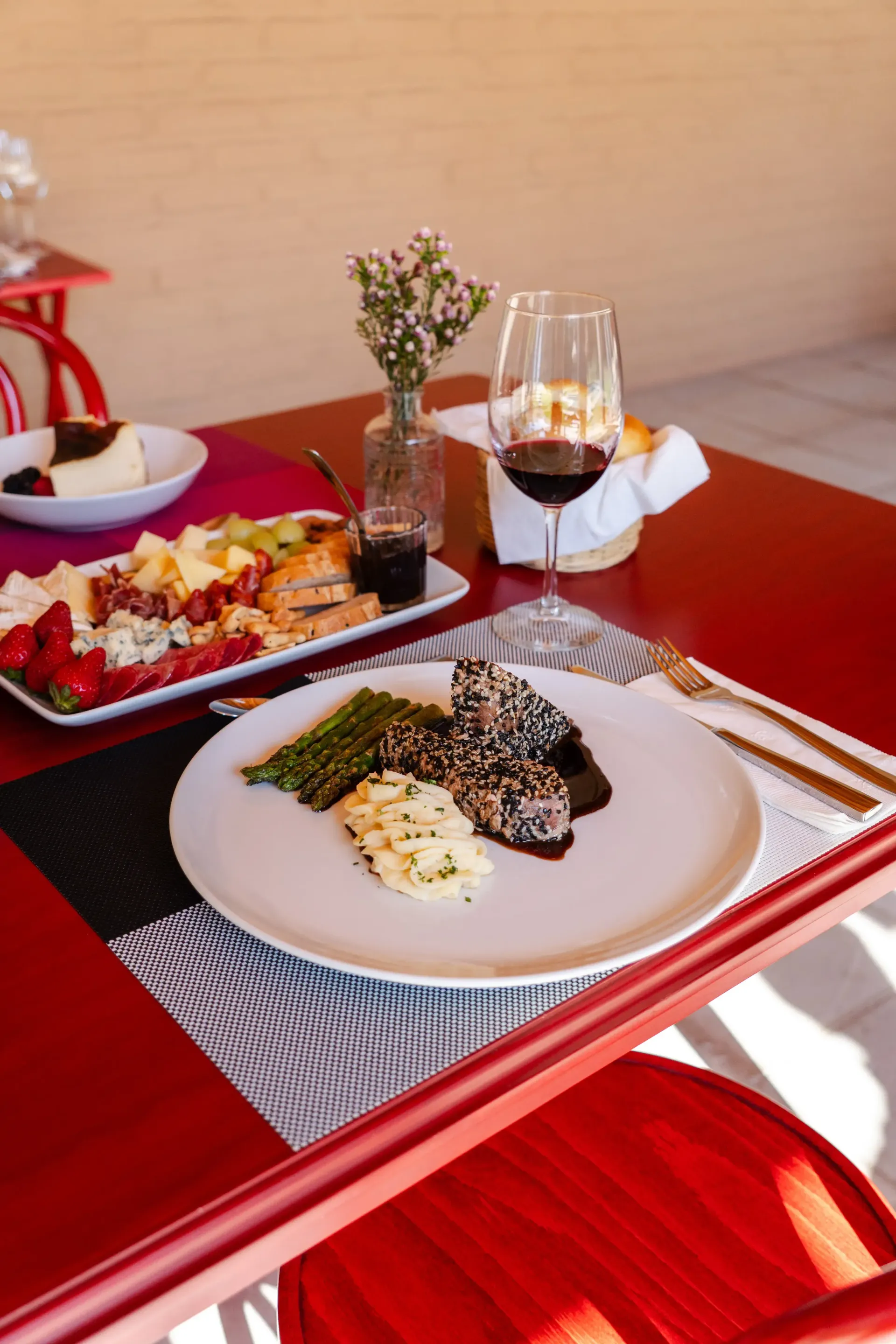 Vino y comida emplatada sobre una mesa de comedor roja, con un pequeño jarrón de flores