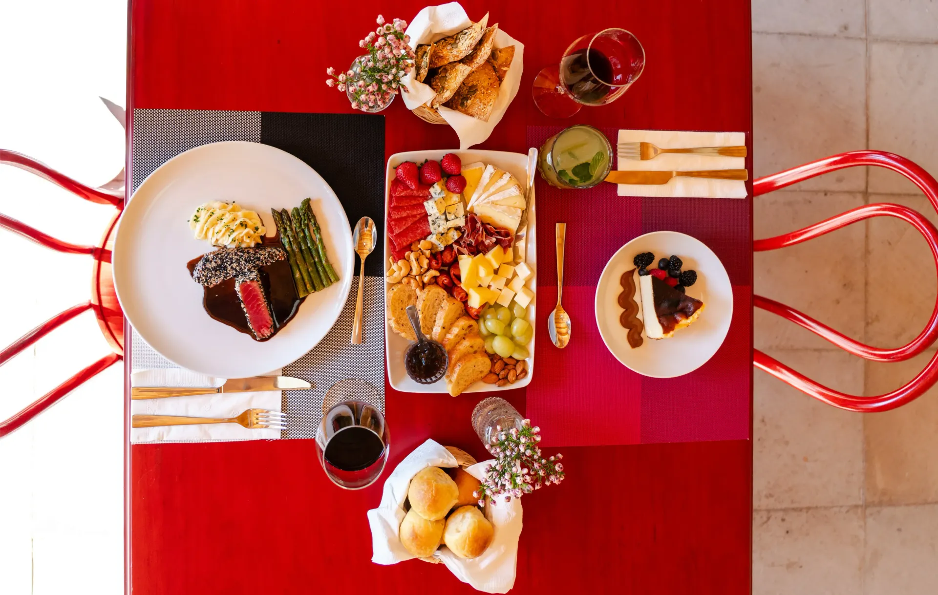 Vista cenital de una mesa roja con postres emplatados, bebidas y pasteles en un patio.