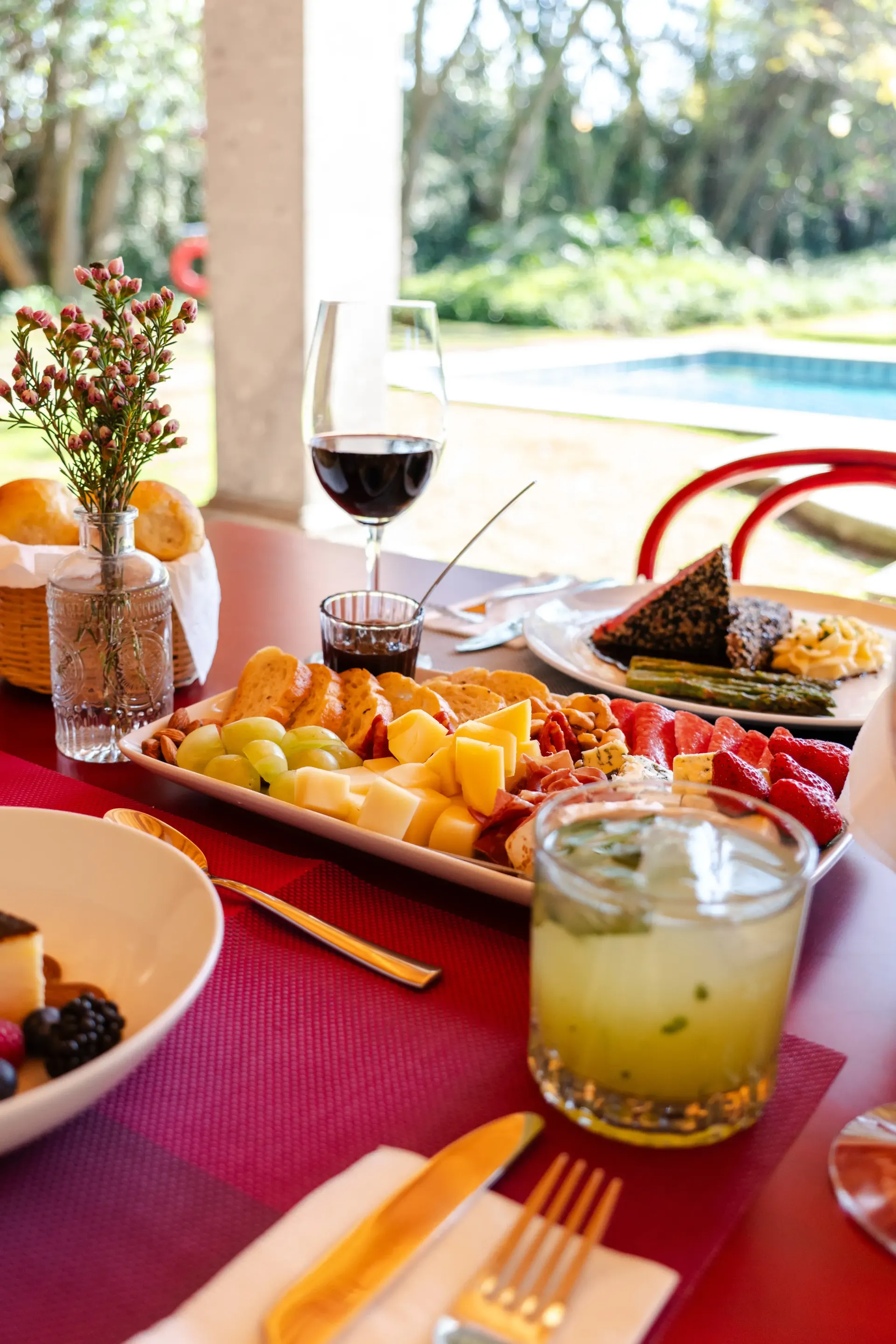 Mesa al aire libre junto a una piscina soleada, con una fuente de frutas, una copa de vino, una ensalada y una bebida