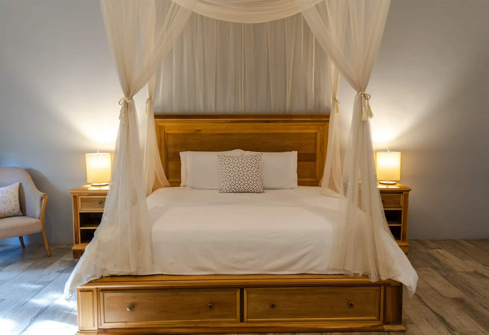 Cama con dosel y cortinas blancas en un dormitorio con iluminación tenue, flanqueada por lámparas de mesita de noche a juego.