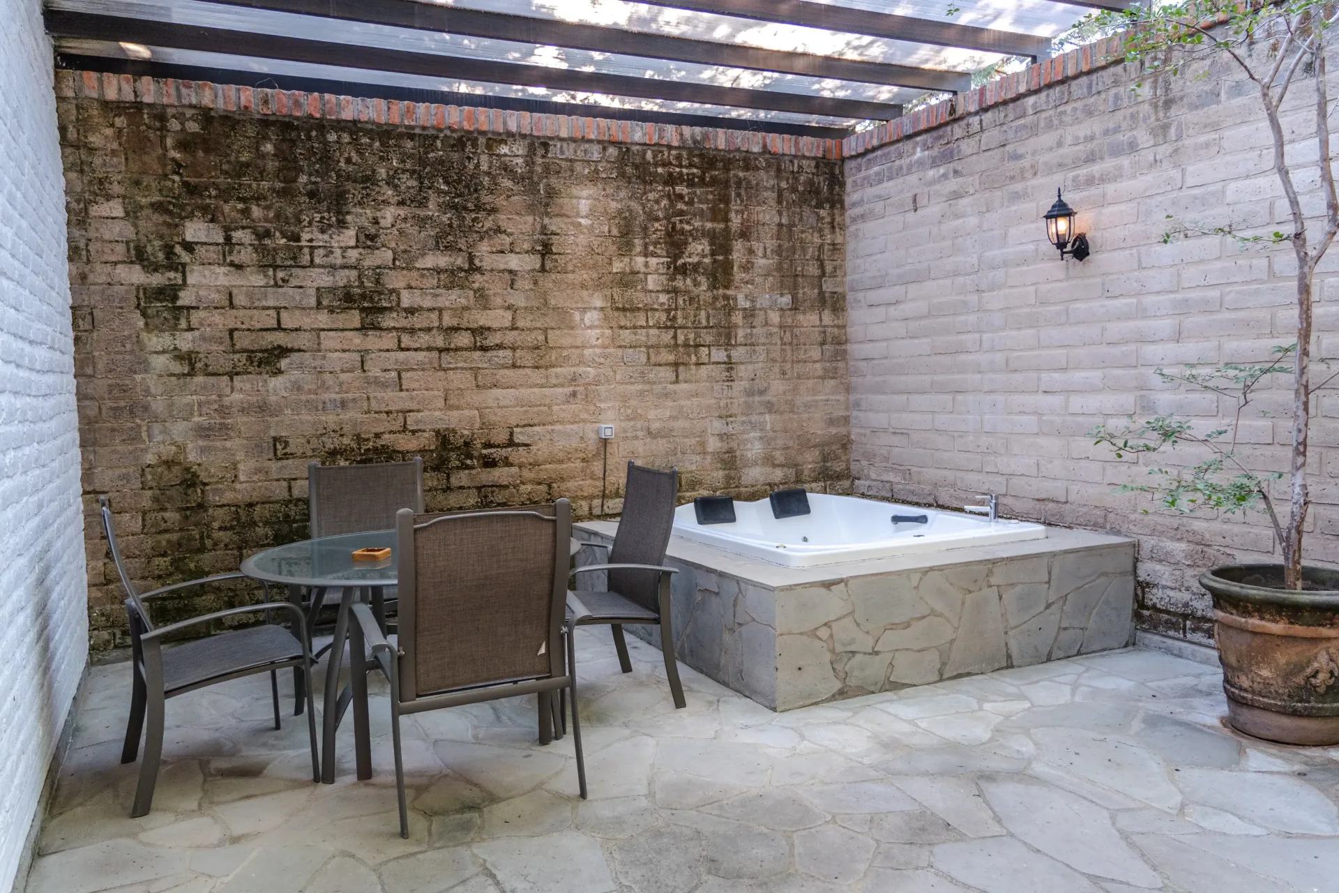 Pequeño patio de piedra con una bañera de hidromasaje, sillas y plantas en macetas bajo un techo de listones