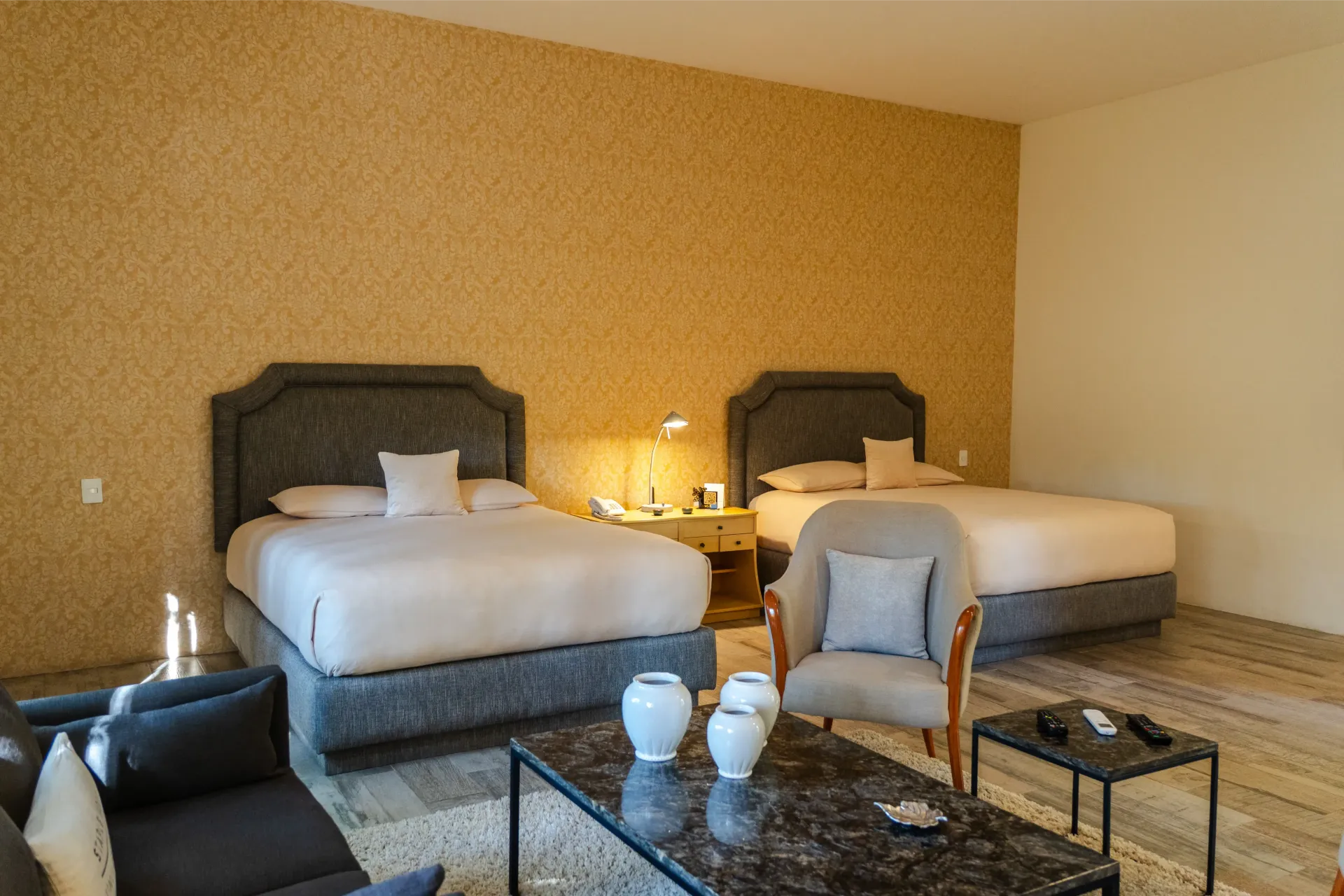 Habitación de hotel con dos camas queen, lámpara de mesita, sillón y mesa de centro de cristal