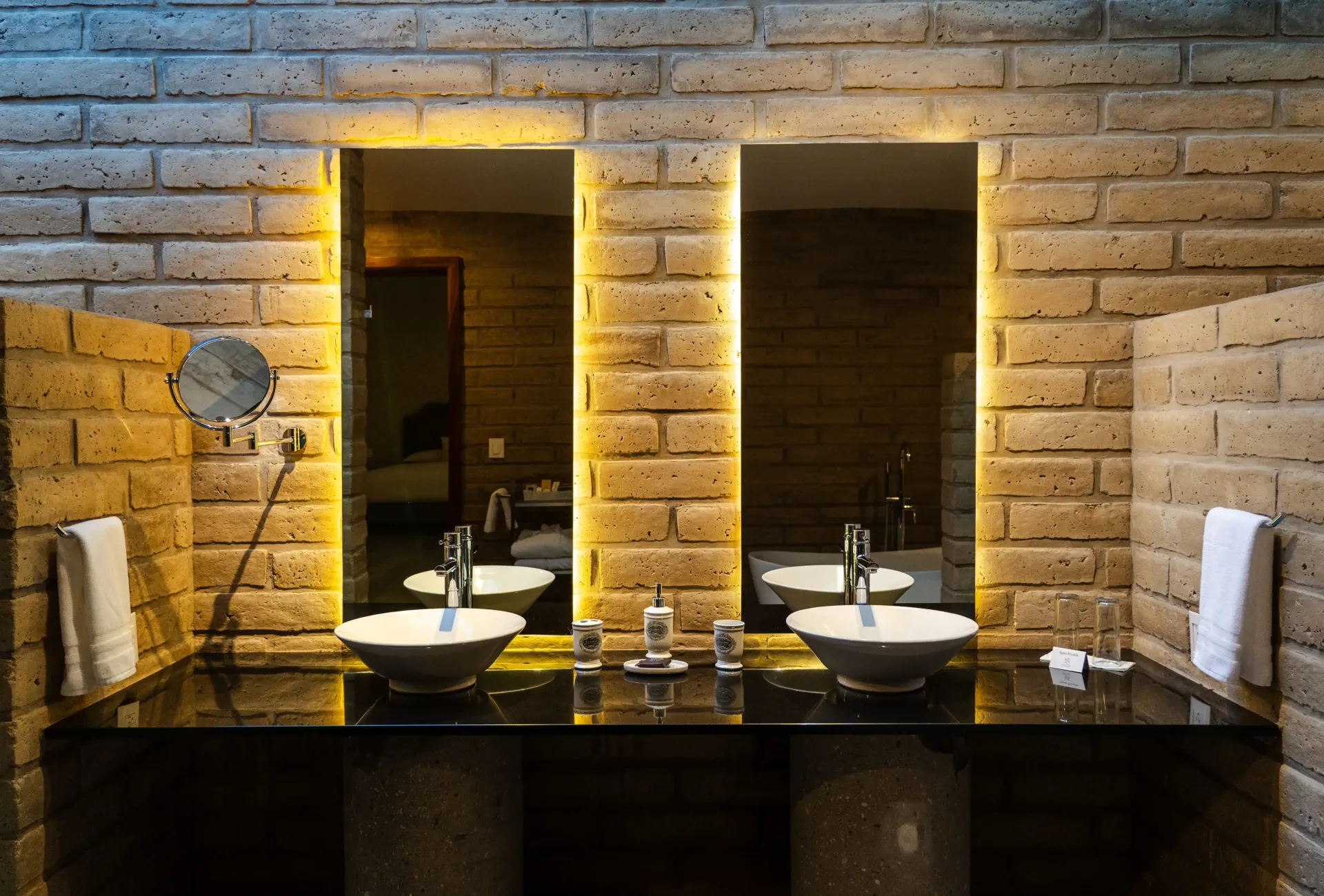 Dos lavabos de baño iluminados con espejos, sobre una pared de ladrillo y una encimera oscura.