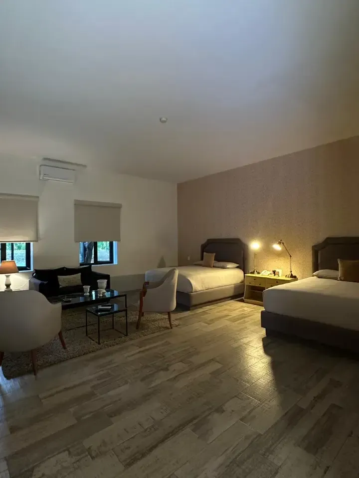 Habitación de hotel con tres camas, una mesa junto a la ventana y una lámpara en la mesita de noche, iluminada con luz cálida por la noche