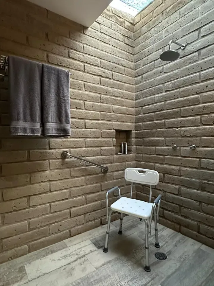 Pequeña ducha alicatada con un taburete blanco, una toalla, una alcachofa de ducha montada en la pared y un desagüe en el suelo.