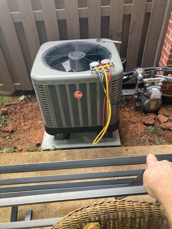 An air conditioning unit with hoses attached, set on a concrete pad near a brick wall and wooden fence.