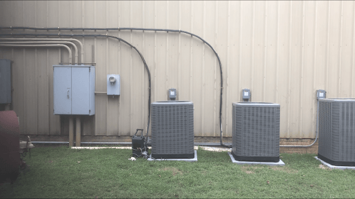 Air conditioning units outside a building with electrical conduit and a panel.