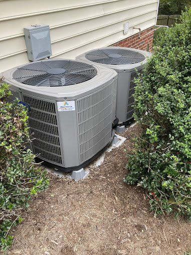 Two gray air conditioning units next to a house, partially obscured by green bushes.