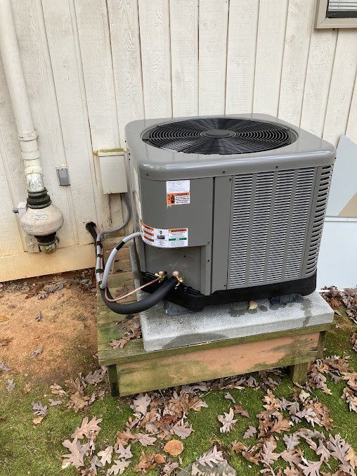An air conditioning unit on a wooden platform beside a white building. Ground covered in leaves.