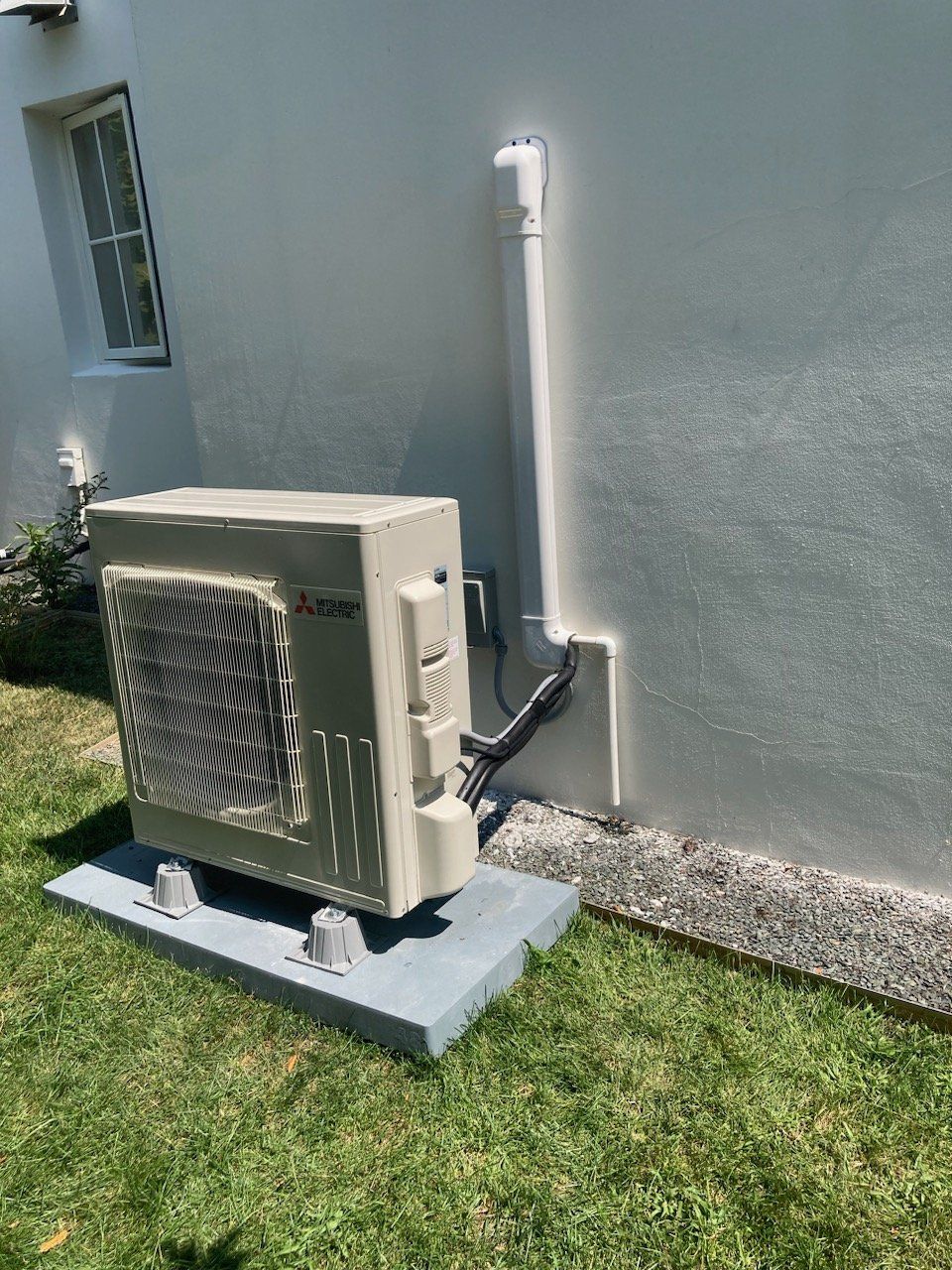 Outdoor air conditioning unit against a white wall; gray base, green grass, and small window in background.