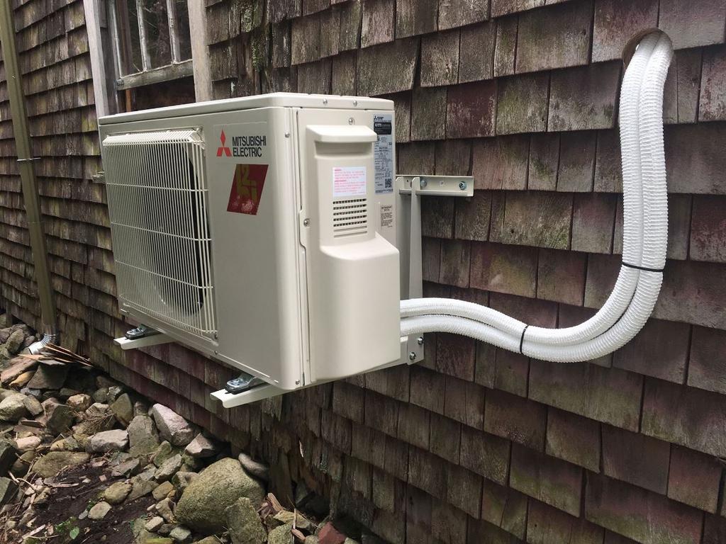 Exterior air conditioning unit mounted on a wood-shingled wall; white tubing runs to the unit.