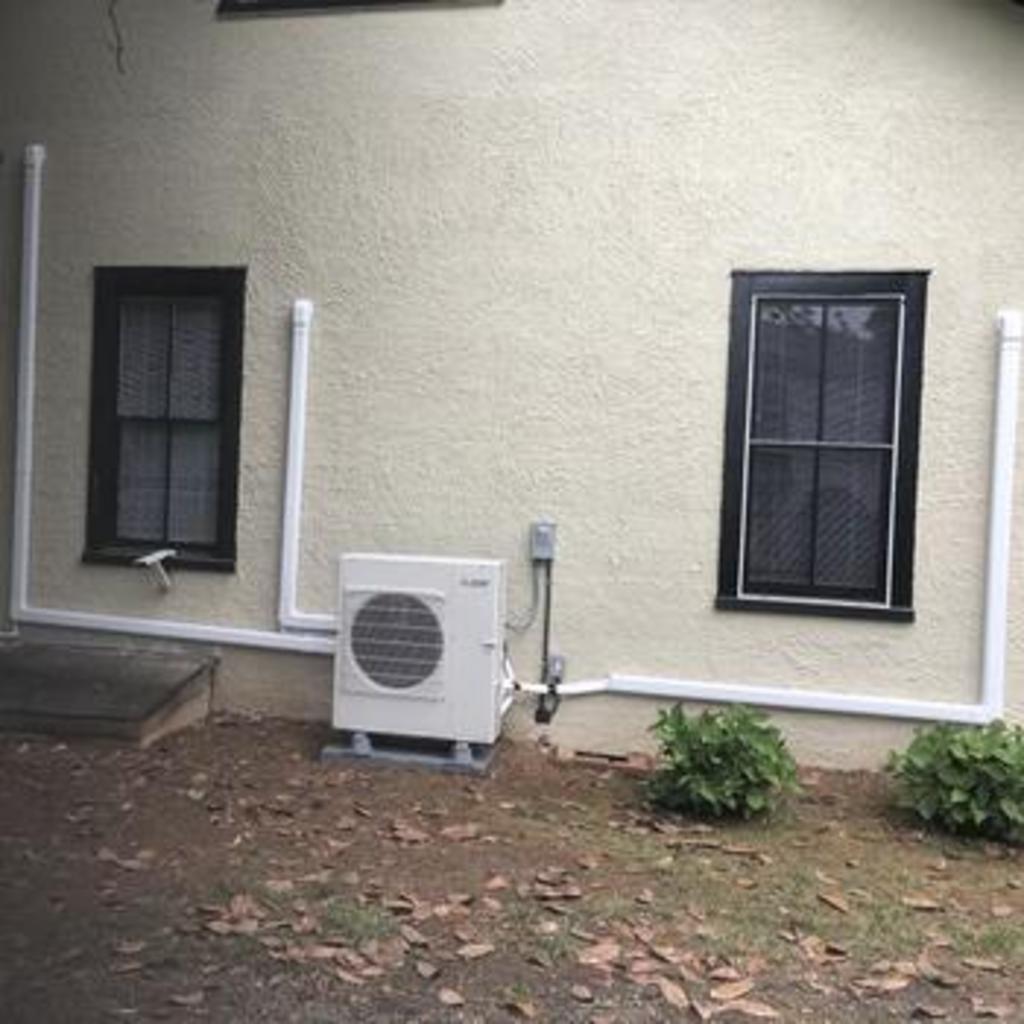 An air conditioning unit on a beige building exterior with two windows and white pipes.