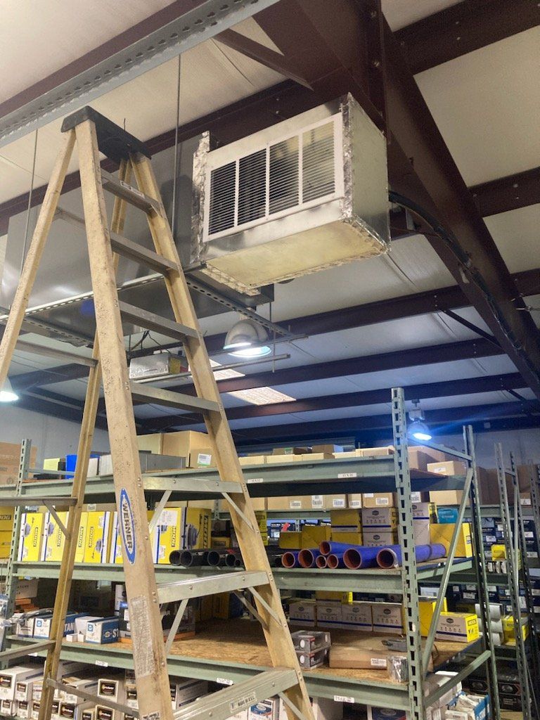 Ladder reaching an overhead ventilation unit in a warehouse with shelving units filled with items.