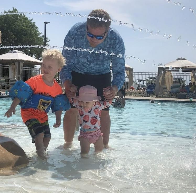 A man helps two young children in a pool; one child wears arm floaties, the other a pink hat.