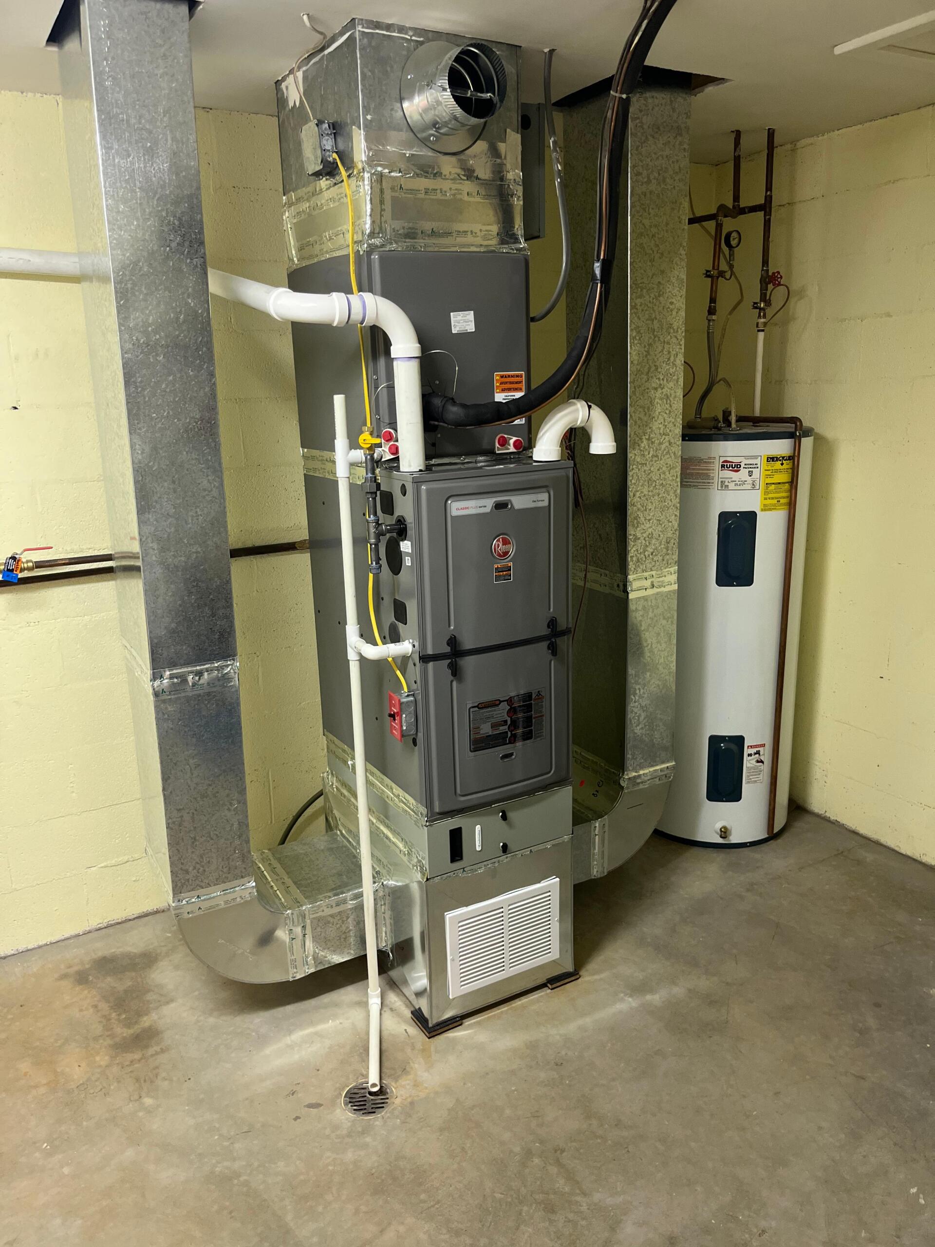 Furnace and water heater in a utility room. Gray metal units with connected ductwork. Beige walls and concrete floor.
