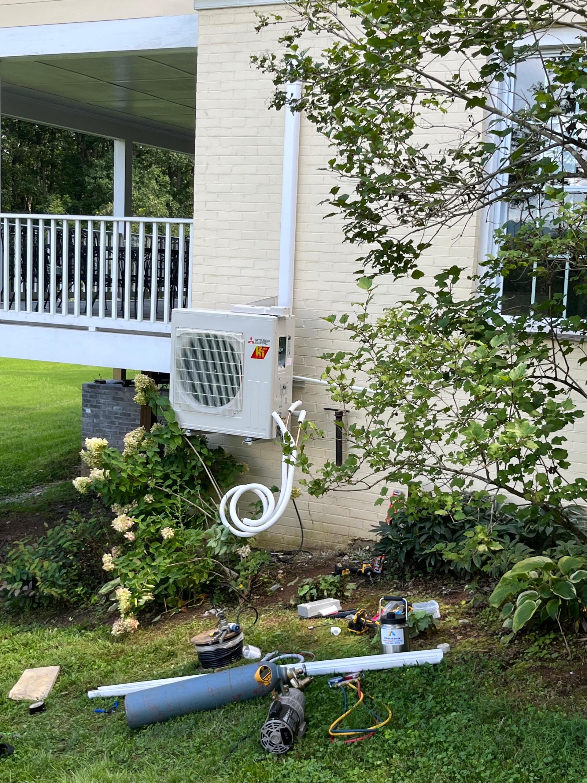 HVAC unit mounted on a beige brick wall with white piping. Ground has tools and supplies on grass.
