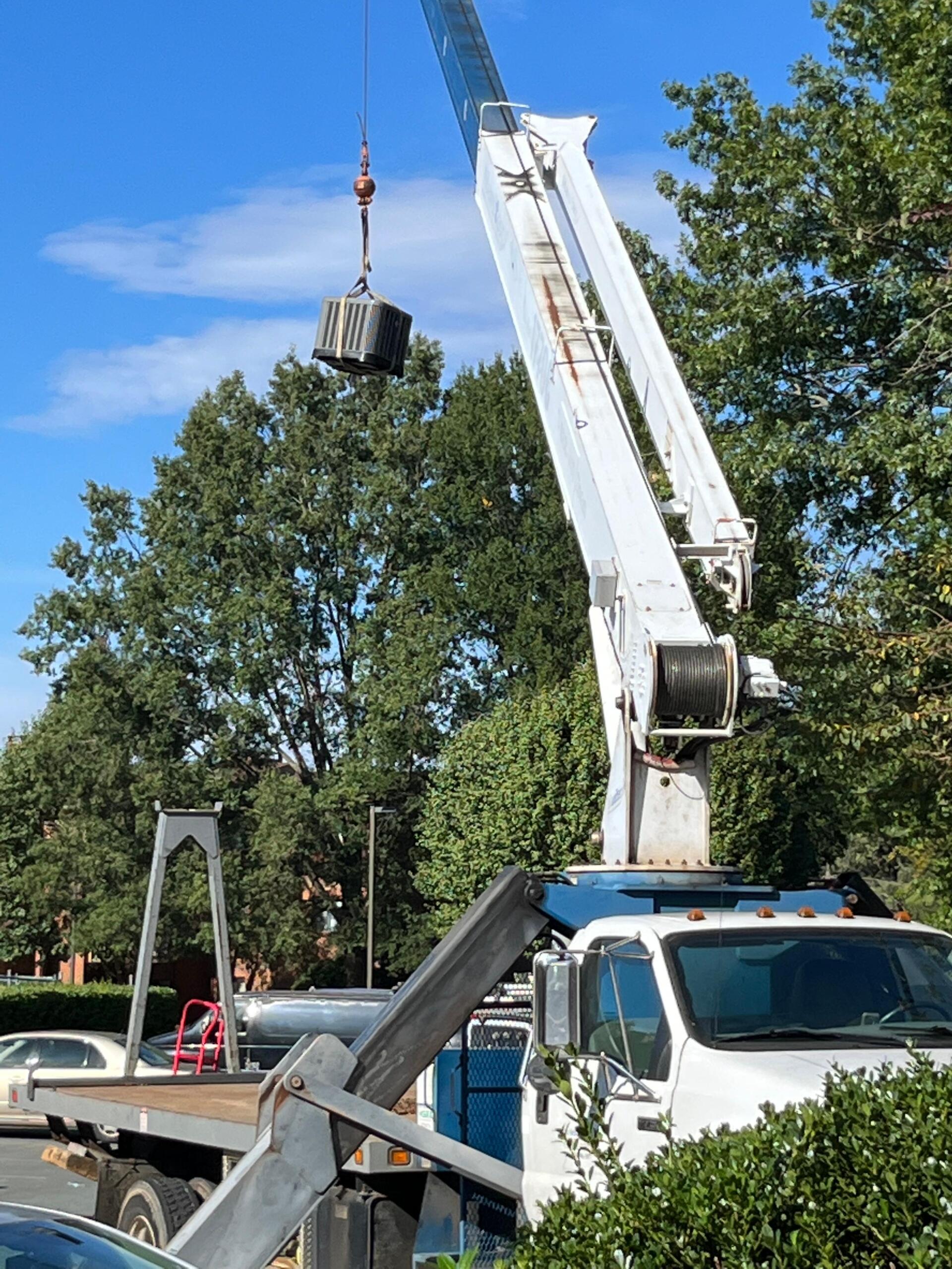 Crane lifting a box in a residential area; blue crane on a white truck. Trees and sky in the background.