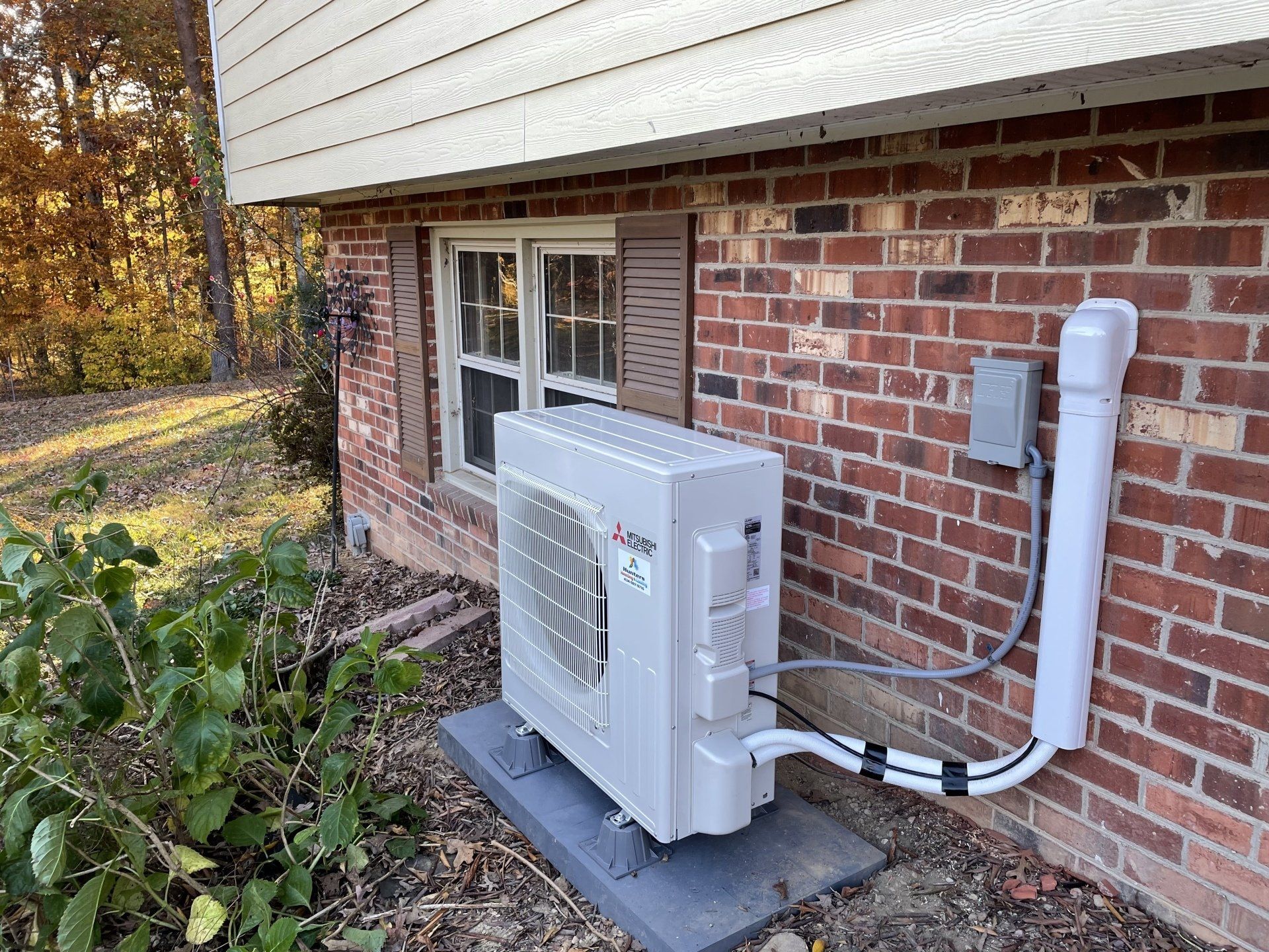 Mini-split heat pump installed on brick exterior wall next to a window and bushes.