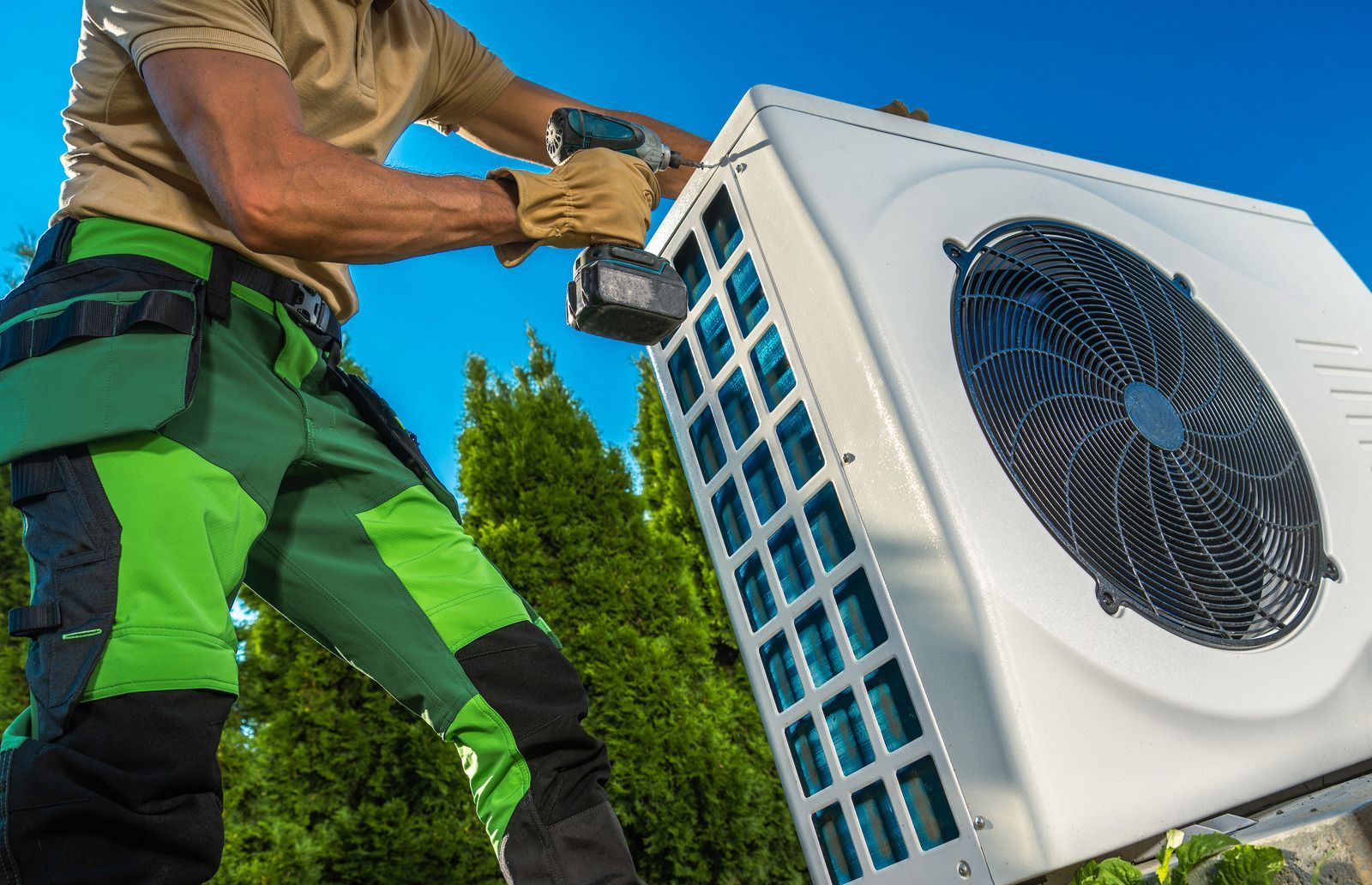 HVAC technician installing an outdoor air conditioning unit with a drill. Bright blue sky background.