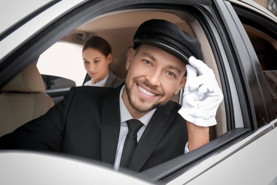 Smiling chauffeur in black suit and cap, tipping his hat from a car window with a passenger behind him.