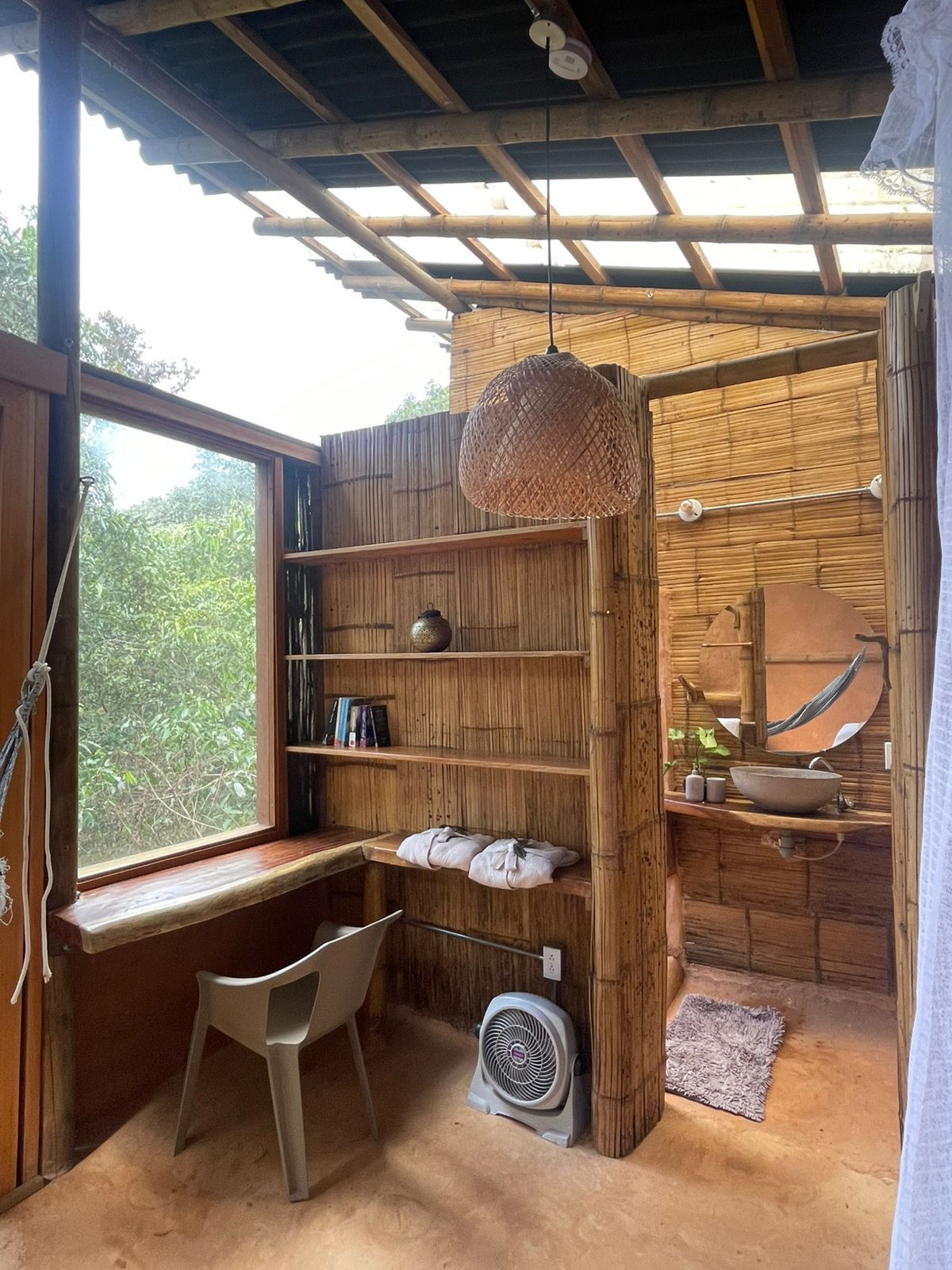 A room with a desk , chair , fan and a window.