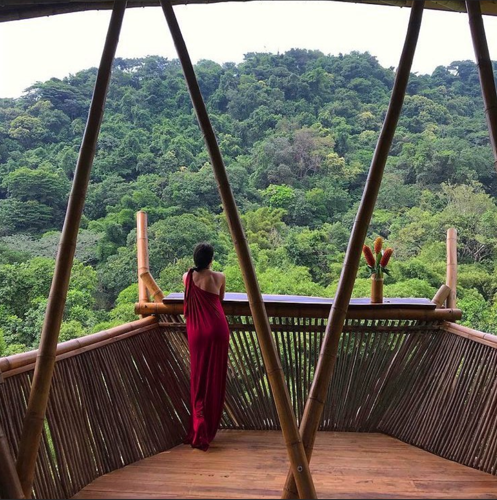 A woman in a red dress stands on a balcony overlooking a forest
