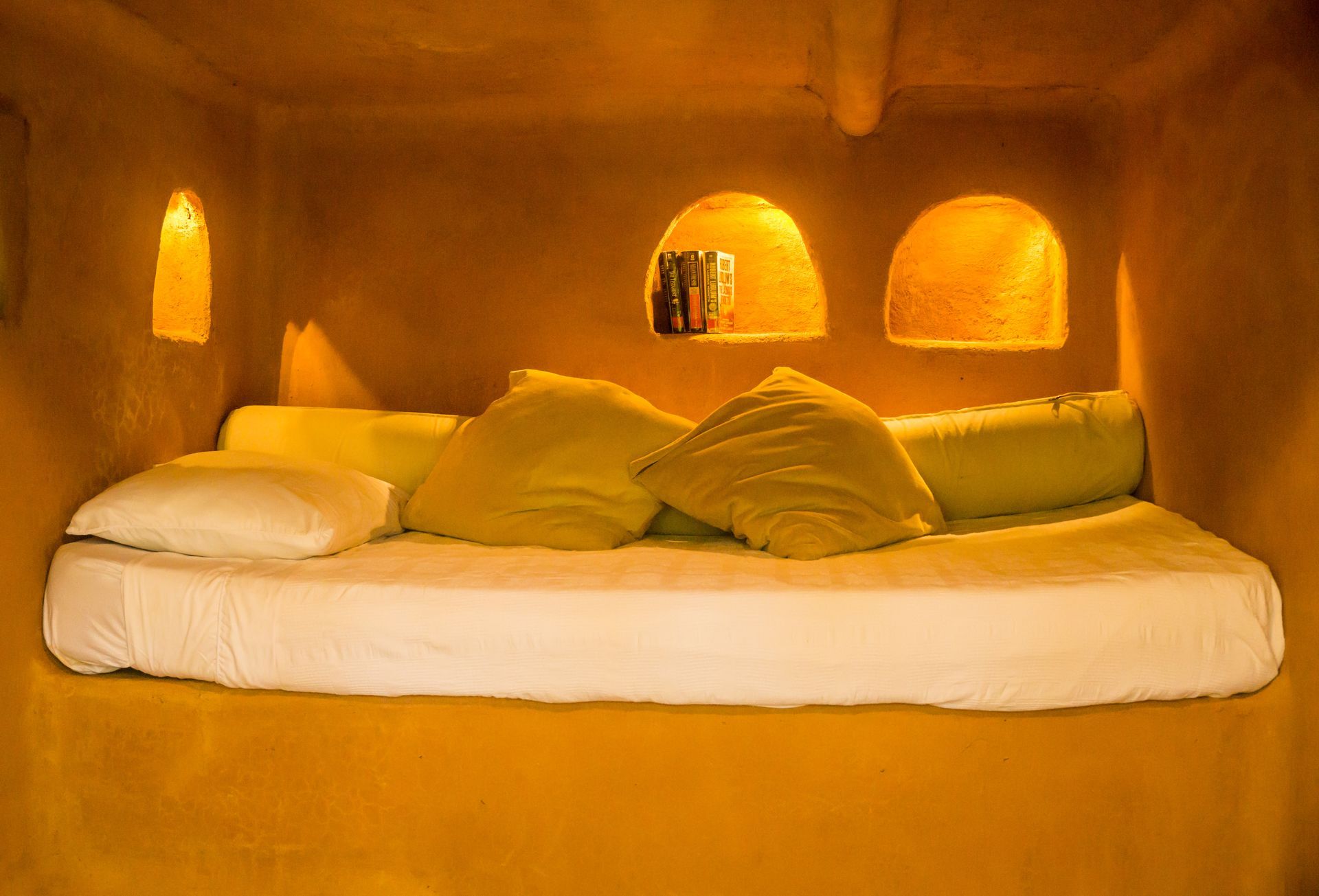 A bed with white sheets and yellow pillows in a room with arches on the wall.
