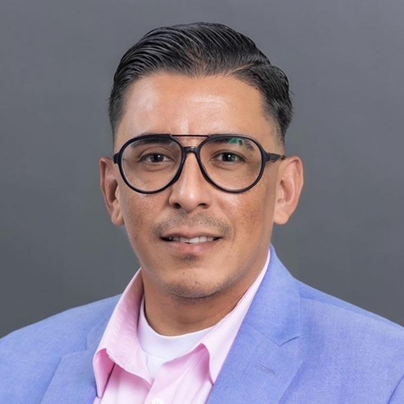 Professional headshot of a man in glasses wearing a light blue blazer and pink shirt against a gray background
