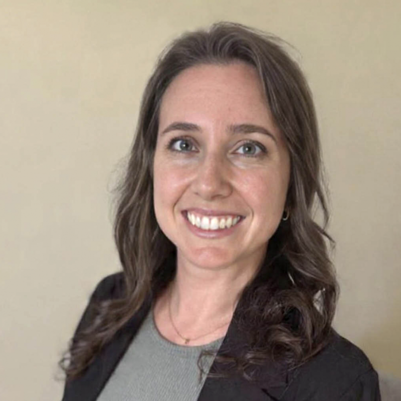 Smiling woman with dark wavy hair in a black blazer against a beige background