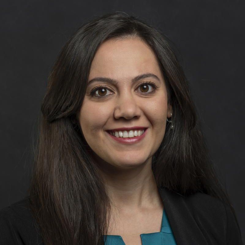 Portrait of a smiling woman with dark hair against a dark background, wearing a black blazer and blue top