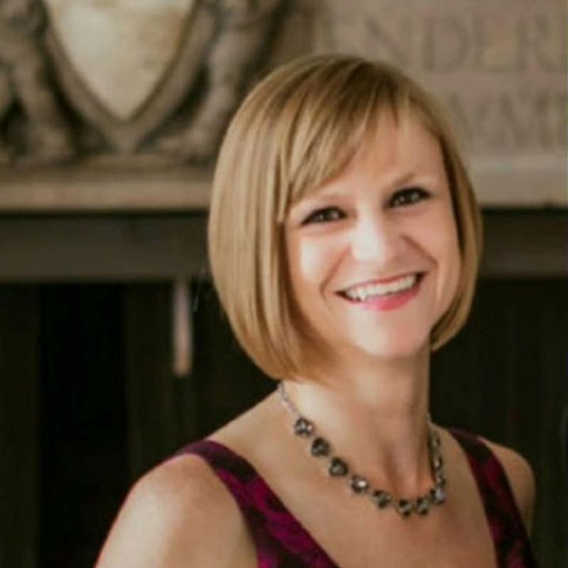 Smiling woman with short blonde hair in a burgundy dress and necklace, standing indoors