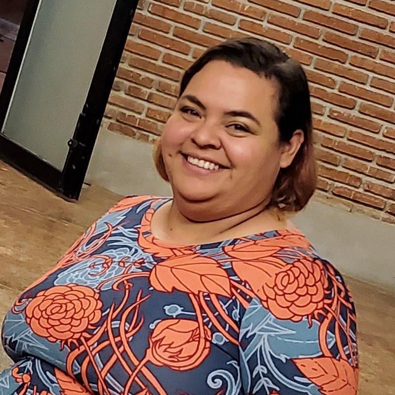 Smiling person in a blue and orange floral top standing outdoors near a brick wall