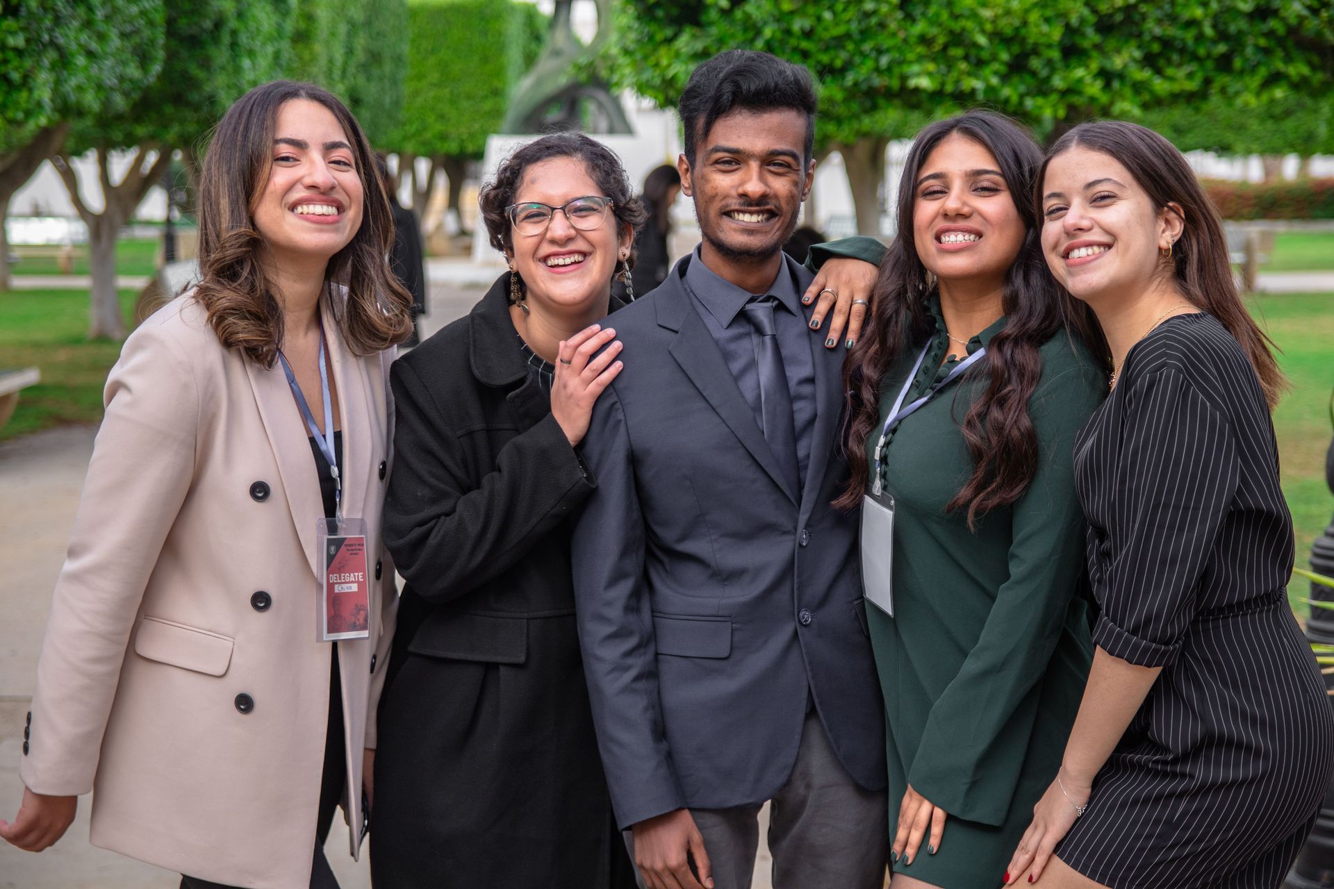 Five smiling people in formal attire standing together outdoors on a campus walkway.