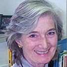 Smiling woman with short gray hair, wearing a light shirt, seated indoors near bookshelves