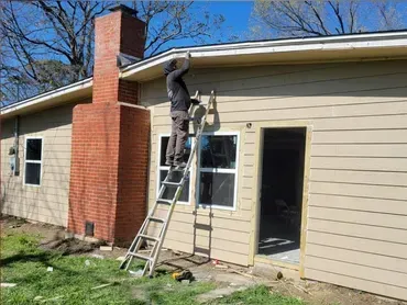 Worker on ladder repairing roof gutter on beige house beside brick chimney.