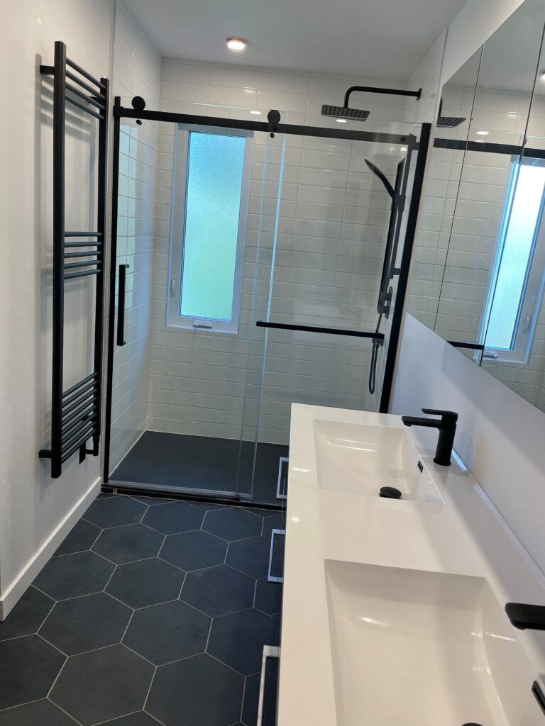 Salle de bains moderne avec sol en carrelage hexagonal foncé, douche en verre, robinetterie noire et meuble-lavabo blanc.