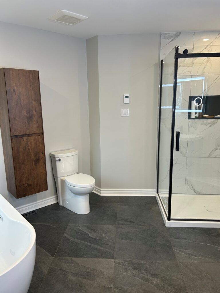 Salle de bains moderne : toilettes, douche avec cadre noir, sol carrelé foncé, armoire haute en bois, murs gris clair.
