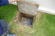 Open utility pit in grass beside a wall, with a person lifting the concrete cover.