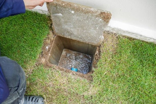 Open utility pit in grass beside a wall, with a person lifting the concrete cover.