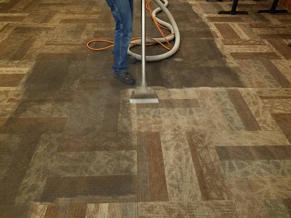 Person vacuuming a patterned carpet with a gray upright vacuum and orange hose