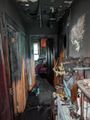 Burned, smoke-damaged hallway with blackened walls and debris, leading to a room with a bed and scattered belongings.