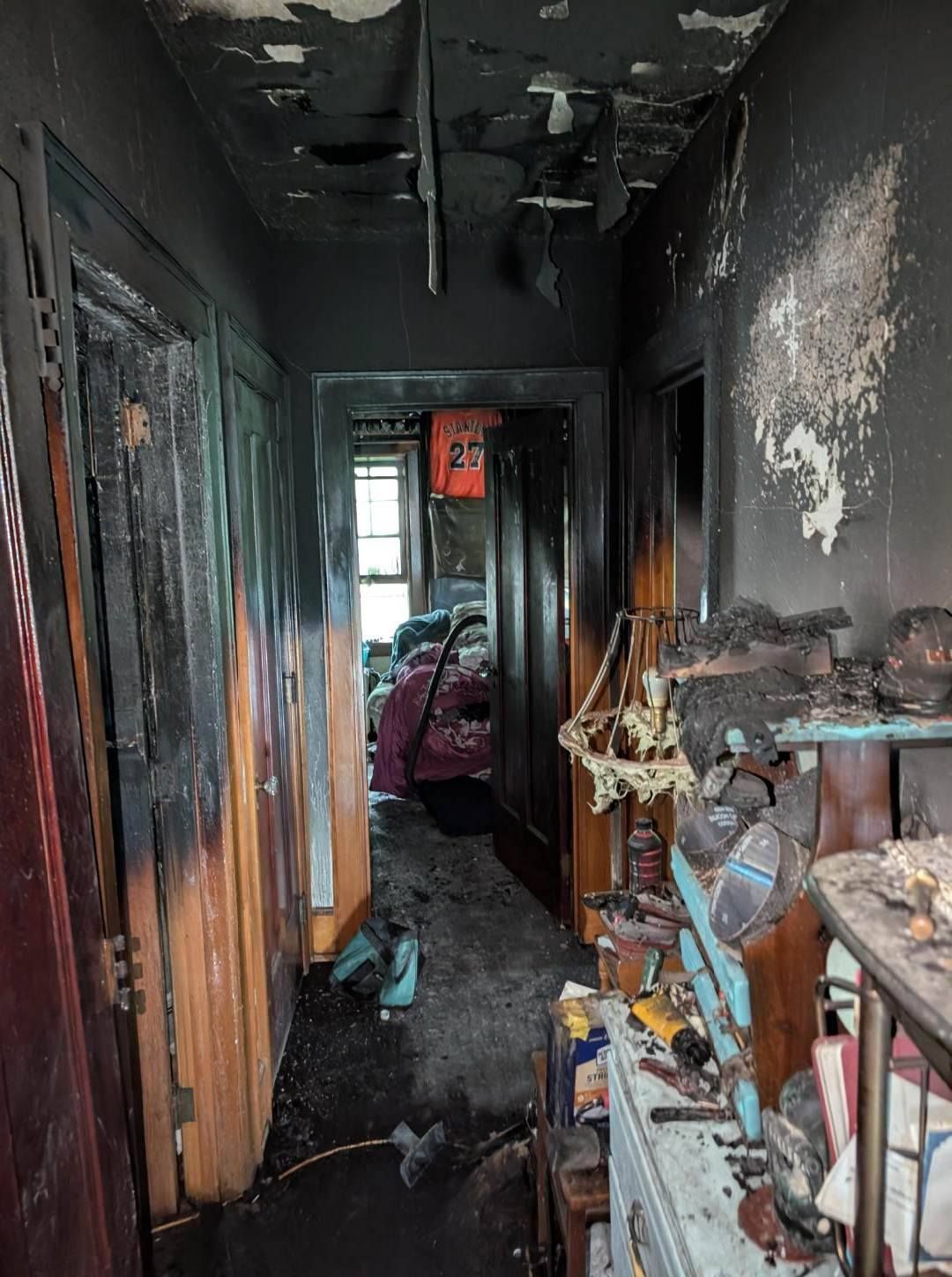 Burned, smoke-damaged hallway with blackened walls and debris, leading to a room with a bed and scattered belongings.